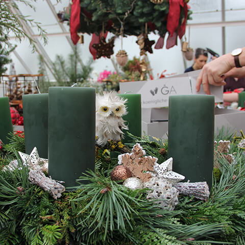 Bei Thea Carlins Do. 27.11.2025 | 10.00-12.00 WORKSHOP - Adventskranzbinden bis zu 30 cm, ein Kranz mit grünen Stumpenkerzen, Tannenzweigen und Ornamenten wird gebastelt, während die Teilnehmer im Hintergrund festliche Dekoration arrangieren.