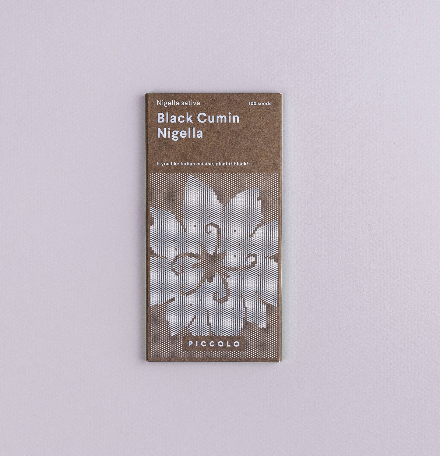 Eine Packung Piccolo Seeds – Nigella Schwarzkümmel liegt auf einer helllavendelfarbenen Oberfläche und zeigt ein weißes Blumenmuster sowie einen Text, der darauf hinweist, dass sie 100 Samen Nigella sativa der Marke Piccolo Seeds enthält.