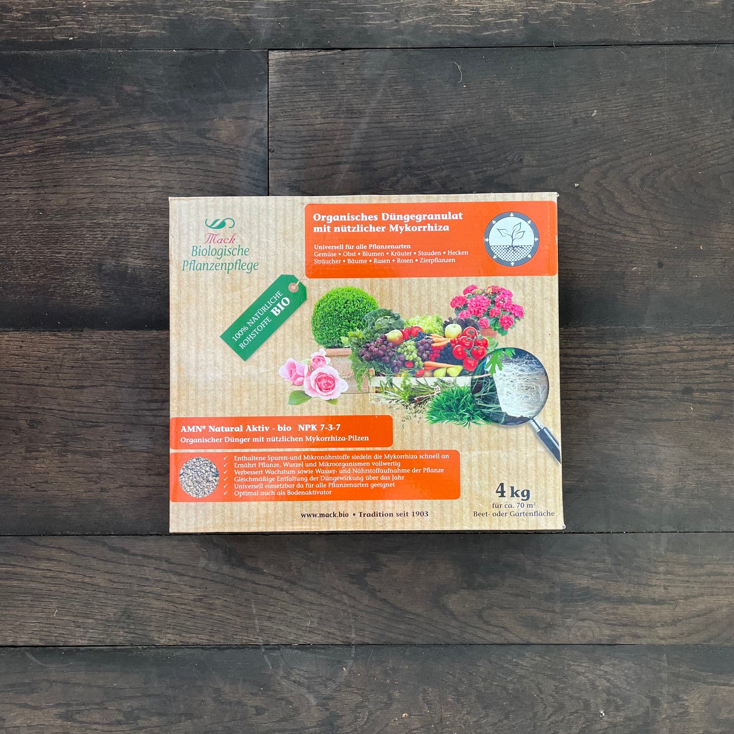 Auf einem dunklen Holzboden steht eine Schachtel AMN Natural Aktiv-bio von AMN. Sie zeigt Bilder von Pflanzen und Blumen sowie einen deutschen Text, der die organischen, Mykorrhiza-reichen Eigenschaften des Produkts hervorhebt.