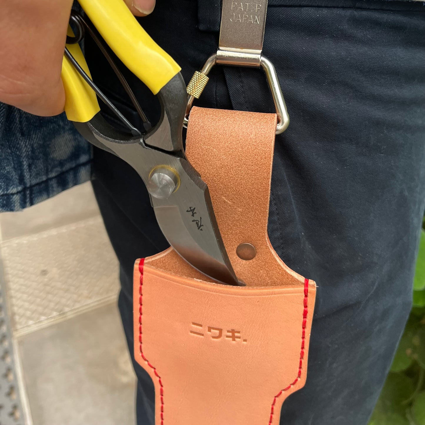 Eine Hand greift nach einer Gartenschere mit gelbem Griff, die an einem Niwaki-Gürtelclip mit japanischer Schrift und roten Nähten befestigt ist, der wiederum an einer Gürtelschlaufe einer dunklen Hose befestigt ist.
