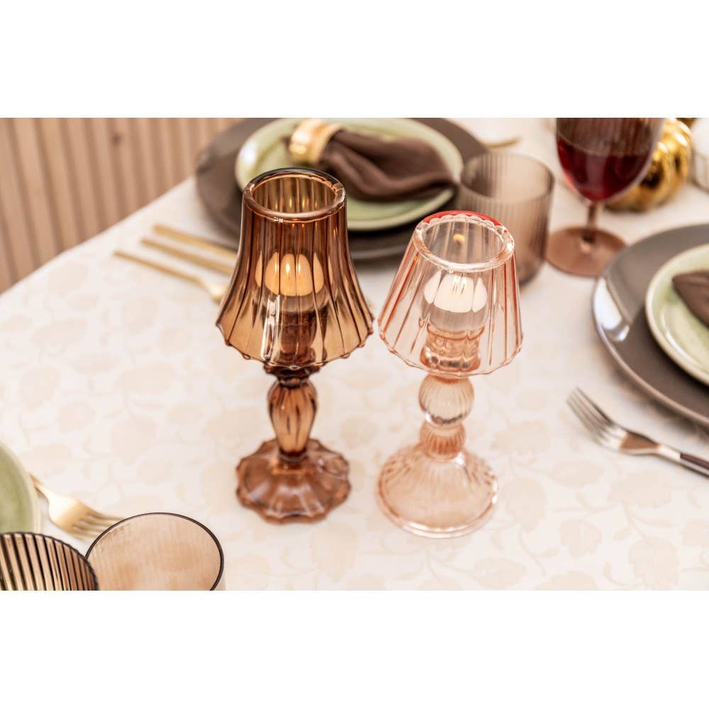 Ein AULICA Brauner Kerzenhalter auf Glasfuß (10x10x24,5cm) steht auf einem weißen Tischtuch neben eleganten Tellern, Servietten, goldenem Besteck und Gläsern.