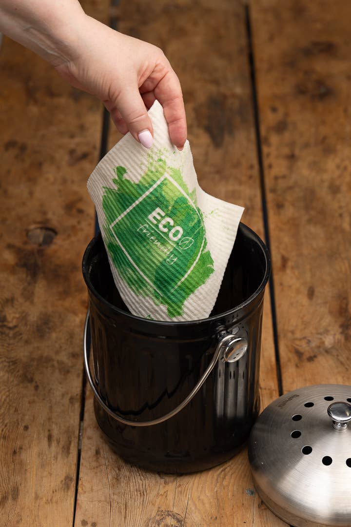 Eine Hand legt ein More Joy - Schwammtuch Appletree von More Joy mit ECO-Label und grüner Blattgrafik in einen schwarzen Komposteimer auf einer Holzoberfläche; der Metalldeckel liegt neben dem Eimer.