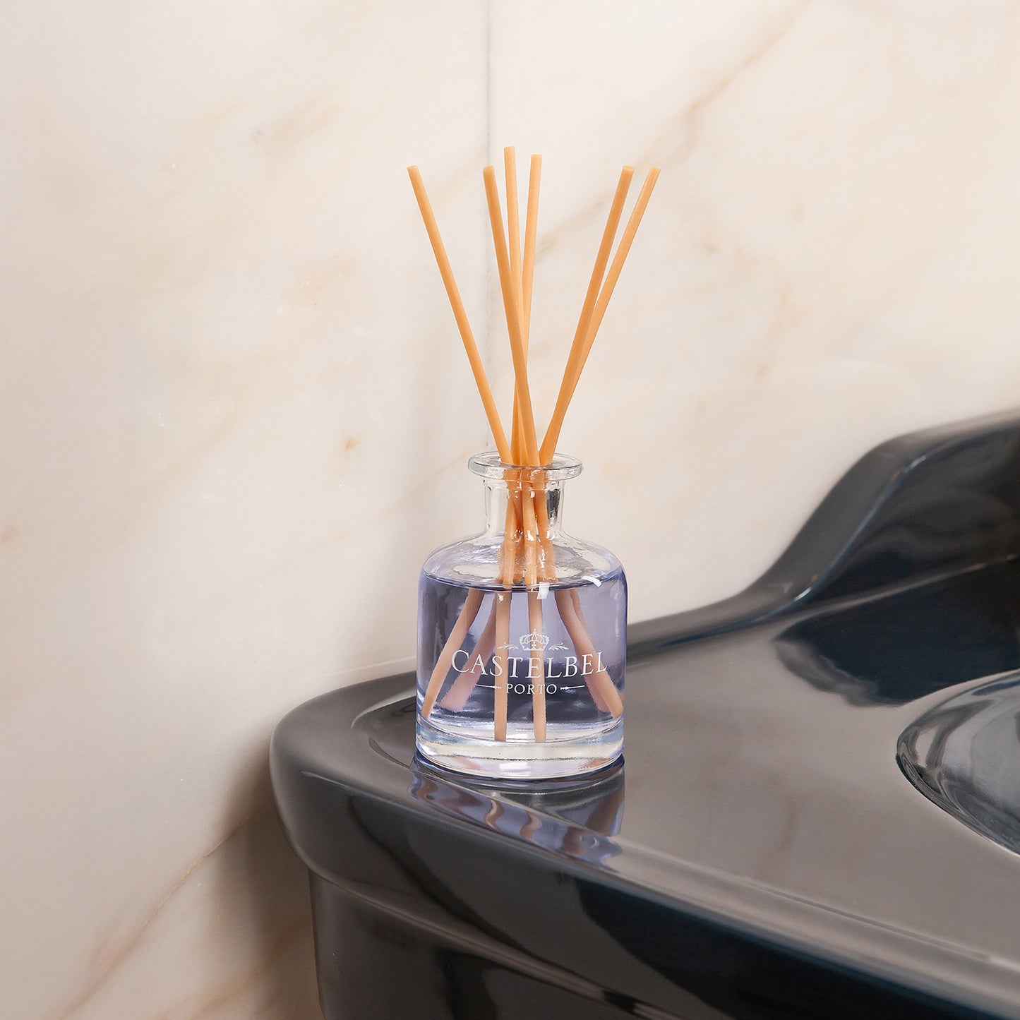 Eine Glasflasche Castelbel Diffusor | Lavendel 250 ml mit violetter Flüssigkeit und Holzstäbchen steht auf einem dunklen Waschbecken vor einer Marmorwand und erfüllt den Raum mit beruhigendem Lavendelduft.