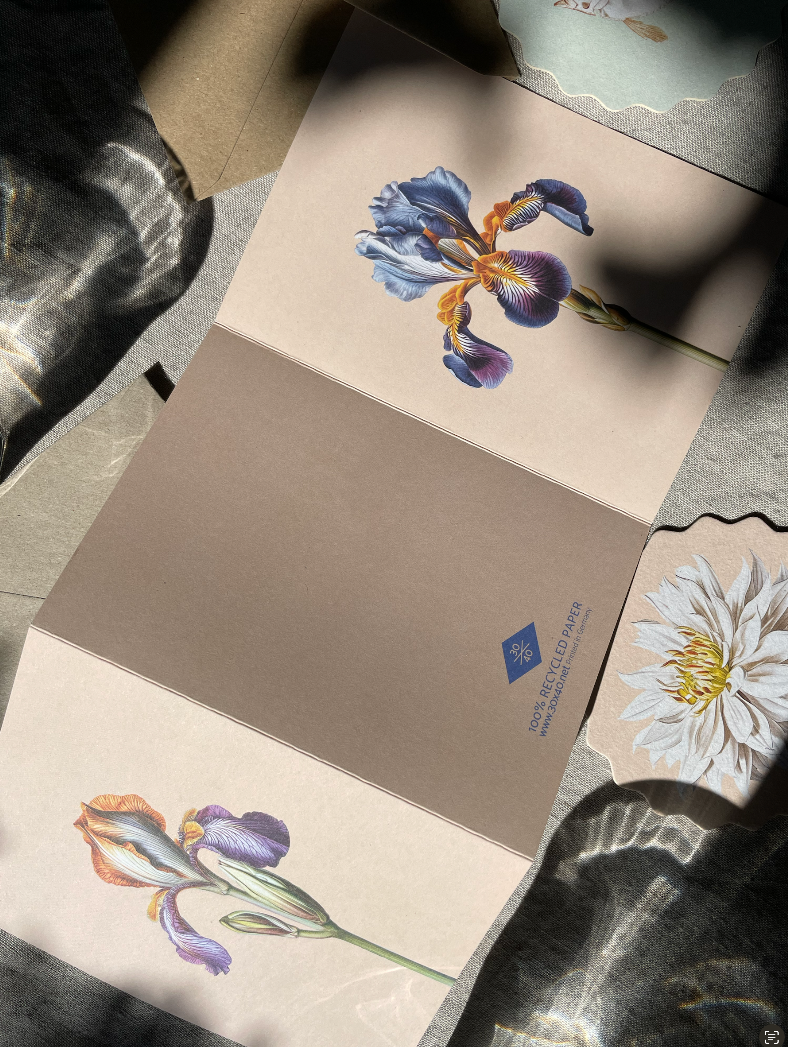 Die in Deutschland auf Recyclingpapier gedruckte 30x40 Iris Leporello Grußkarte von 30x40 zeigt lila Schwertlilien und "Copenhagen 2023" auf Braun. Sie ruht auf einem Stoff mit Wasserspiegelungen; ein floraler Untersetzer mit einer weißen Blüte liegt daneben.