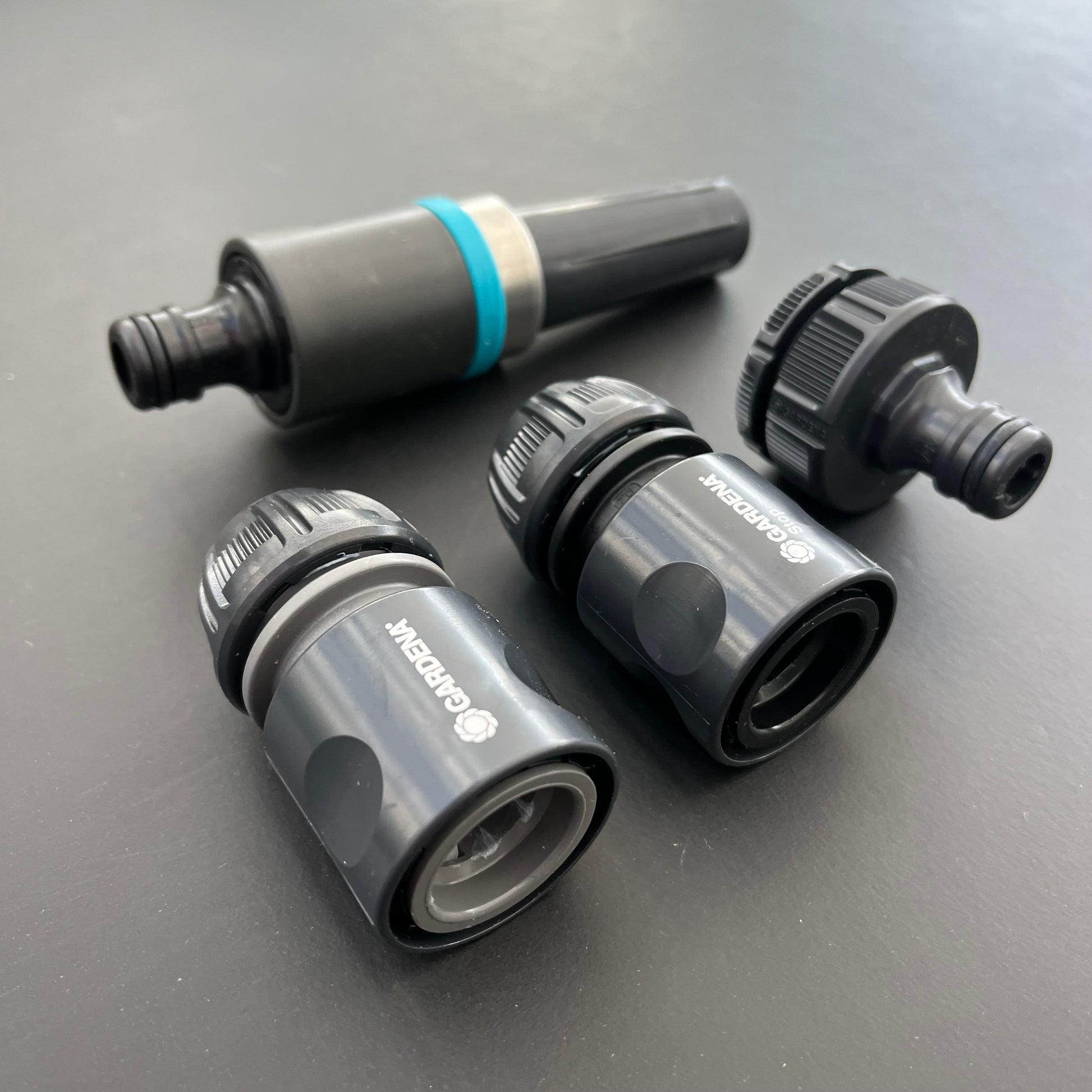 Auf dunklem Untergrund sind drei schwarz-graue Schlauchverbinder und eine Sprühdüse der Gardena EcoLine Grundausstattung abgebildet und heben so die umweltfreundliche Gartenpflege und die langlebigen Materialien der Marke hervor.