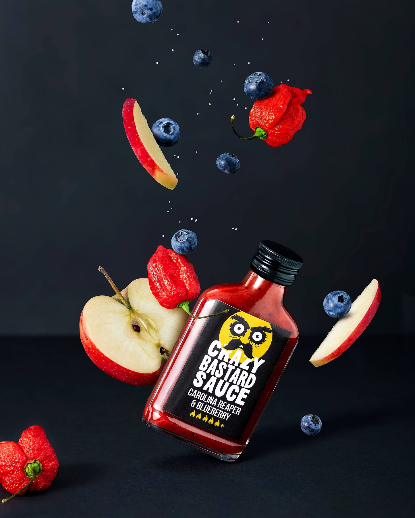 Eine Flasche CBS Foods GmbH (Crazy Bastard Sauce) - Carolina Reaper & Blueberry 100 ml scharfe Soße ist umgeben von schwimmenden Apfelscheiben, feurigen Carolina Reaper Chilis und Blaubeeren, mit Salz oder Gewürzkörnern auf dunklem Grund.