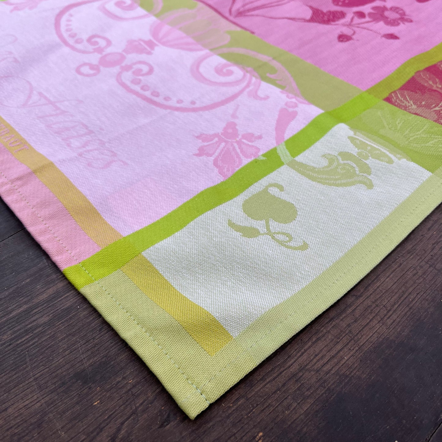 Eine Nahaufnahme des Garnier Thiebaut - Küchenhandtuchs Fraisier, einem Baumwoll-Küchenhandtuch mit genähtem Saum, auf einer dunklen Holzoberfläche. Das Design zeigt rosa und grüne Blumen- und Ornamentmuster in verschiedenen Farbtönen.