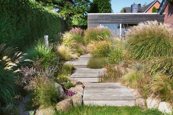 Ein Weg aus breiten hölzernen Trittsteinen schlängelt sich durch Gräser und Blumen und führt zu einem modernen Holzhaus - die perfekte Inspiration für Ihren Traumgarten aus "Die schönsten Terrassen und Sitzplätze" vom Callwey Verlag.