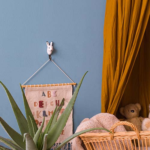 Ein gemütliches Kinderzimmer mit blauer Wand, senfgelbem Vorhang, Korb mit Teddybär und Decken, grüner Aloe-Pflanze und einem Alphabet-Wandbehang an einem Wildlife Garden Handgeschnitzter Wandhaken aus Holz | Schneehase.
