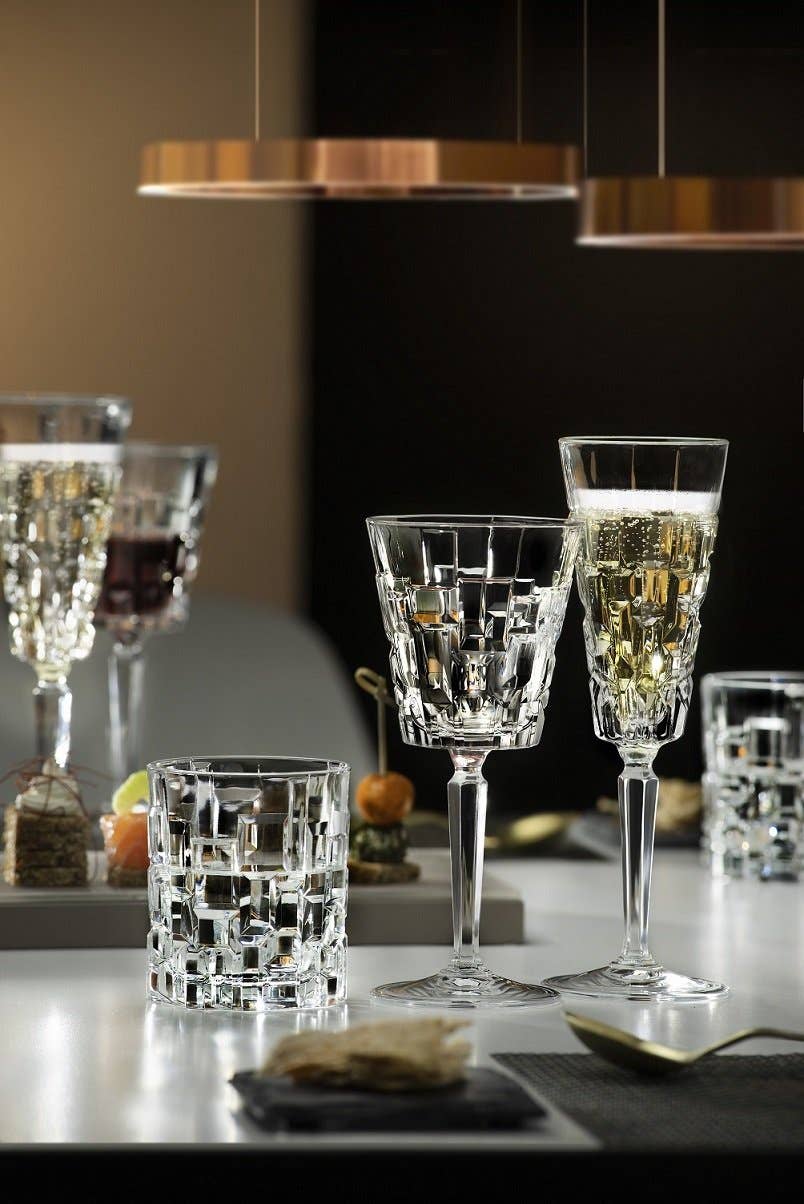 Zwei elegante, mit Weißwein gefüllte Kristallgläser und ein RCR Cristalleria - Tumbler 33 Cl Etna von RCR Cristalleria stehen auf einem glänzenden Tisch und schimmern in ihren Reflexen. Im Hintergrund vervollständigen verschwommene Lichter, Rotwein und Gourmet-Snacks die Szene.
