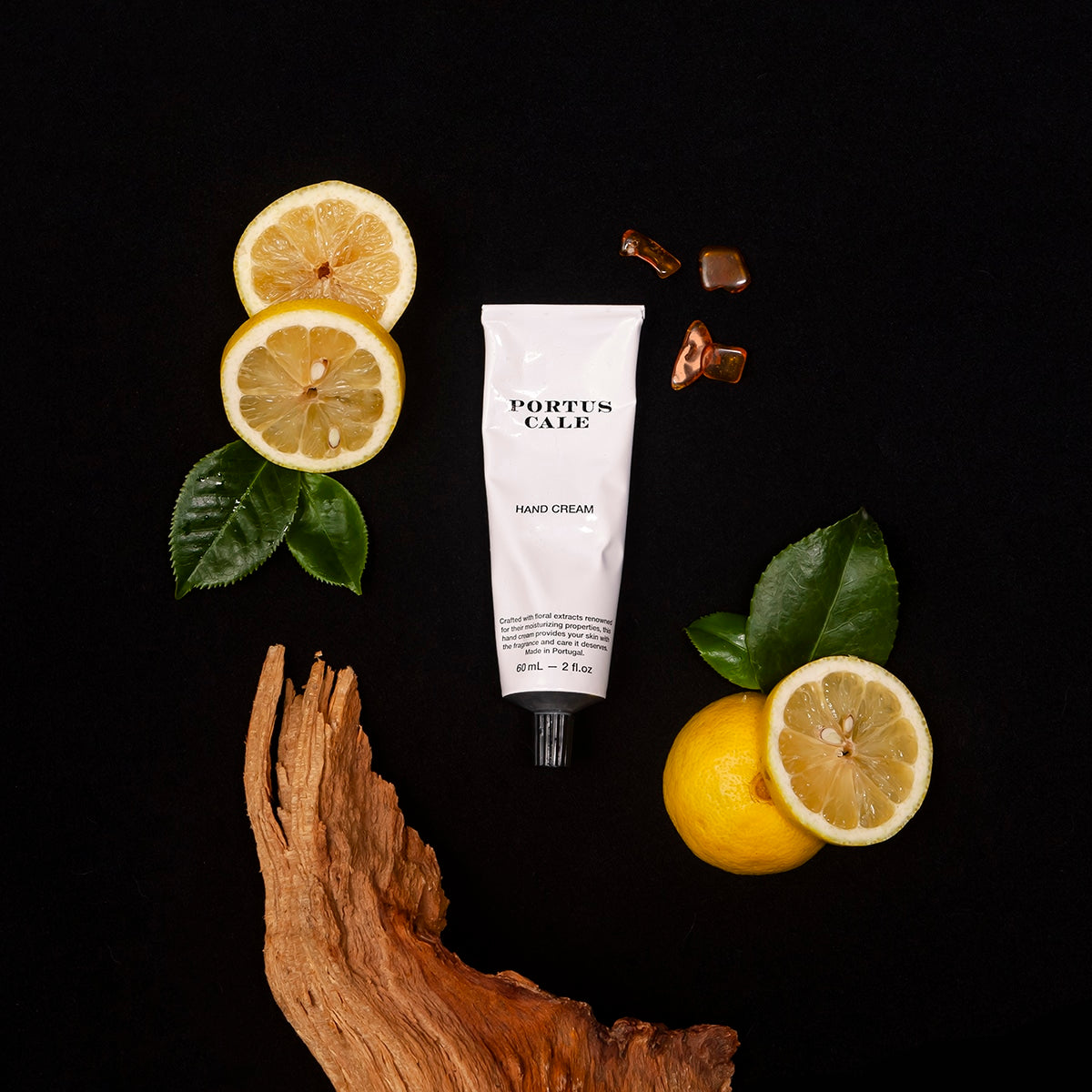 Eine 60-ml-Tube der Handcreme Portus Cale Black Edition von Castelbel ist mit Zitronenscheiben, grünen Blättern, Karamellstücken und Holz verziert und unterstreicht den maskulinen Duft vor einem schlichten schwarzen Hintergrund.