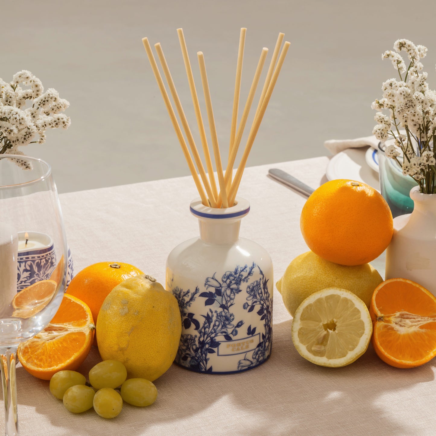 Ein Tisch mit einem Castelbel Diffusor | Gold & Blue 250 ml, umgeben von Orangen, Zitronen, Trauben, weißen Blumen in Vasen, einem Glas und einem Teller auf einer beigen Tischdecke.