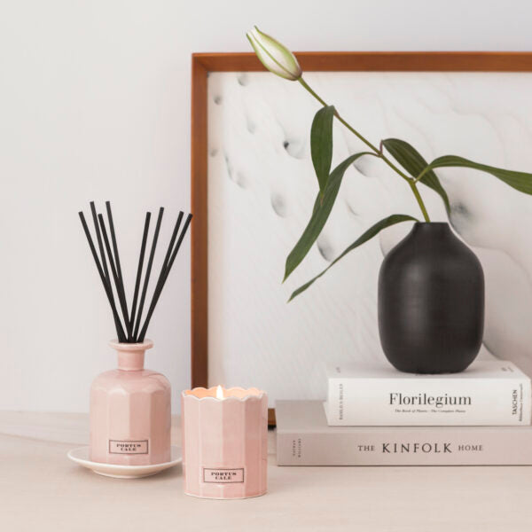 Eine minimalistische Einrichtung mit dem Castelbel Diffusor | Rosé Blush 250 ml, einer brennenden rosa Kerze, einer schwarzen Keramikvase mit einer Lilie und zwei gestapelten Büchern auf einer weißen Fläche vor einem gerahmten Kunstwerk.