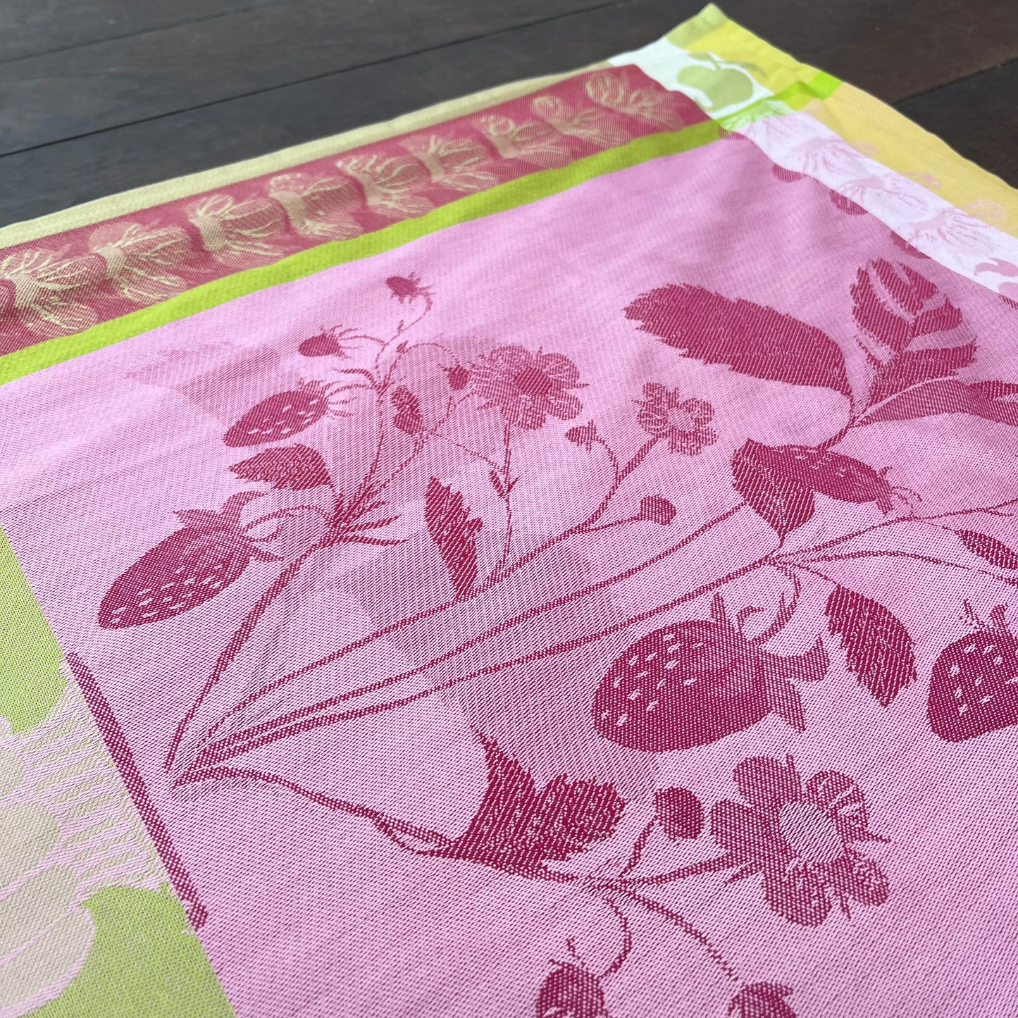 Eine Nahaufnahme des Garnier Thiebaut - Küchenhandtuch Fraisier, ein rosa Küchenhandtuch aus Baumwolle mit eingewebtem Erdbeer- und Blättermuster sowie grünen, gelben und roten Zierrändern, abgebildet auf dunkelbraunem Holz.