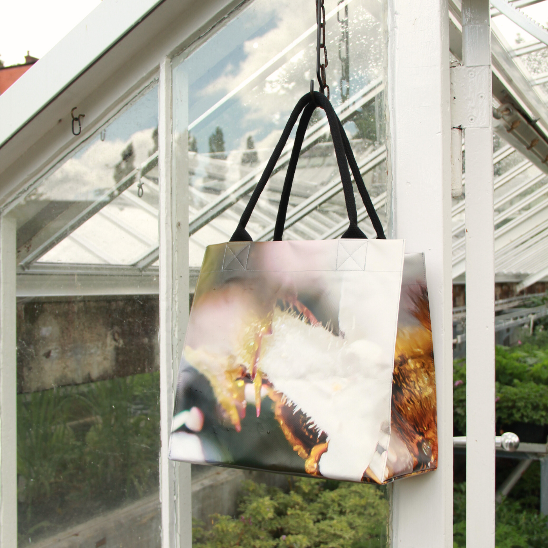 Die Upcycling KGA Tasche 'Biene' von KGA mit einem Naturmotiv in Nahaufnahme hängt an einem Haken in einem Gewächshaus mit Glaswänden und Grünpflanzen draußen.