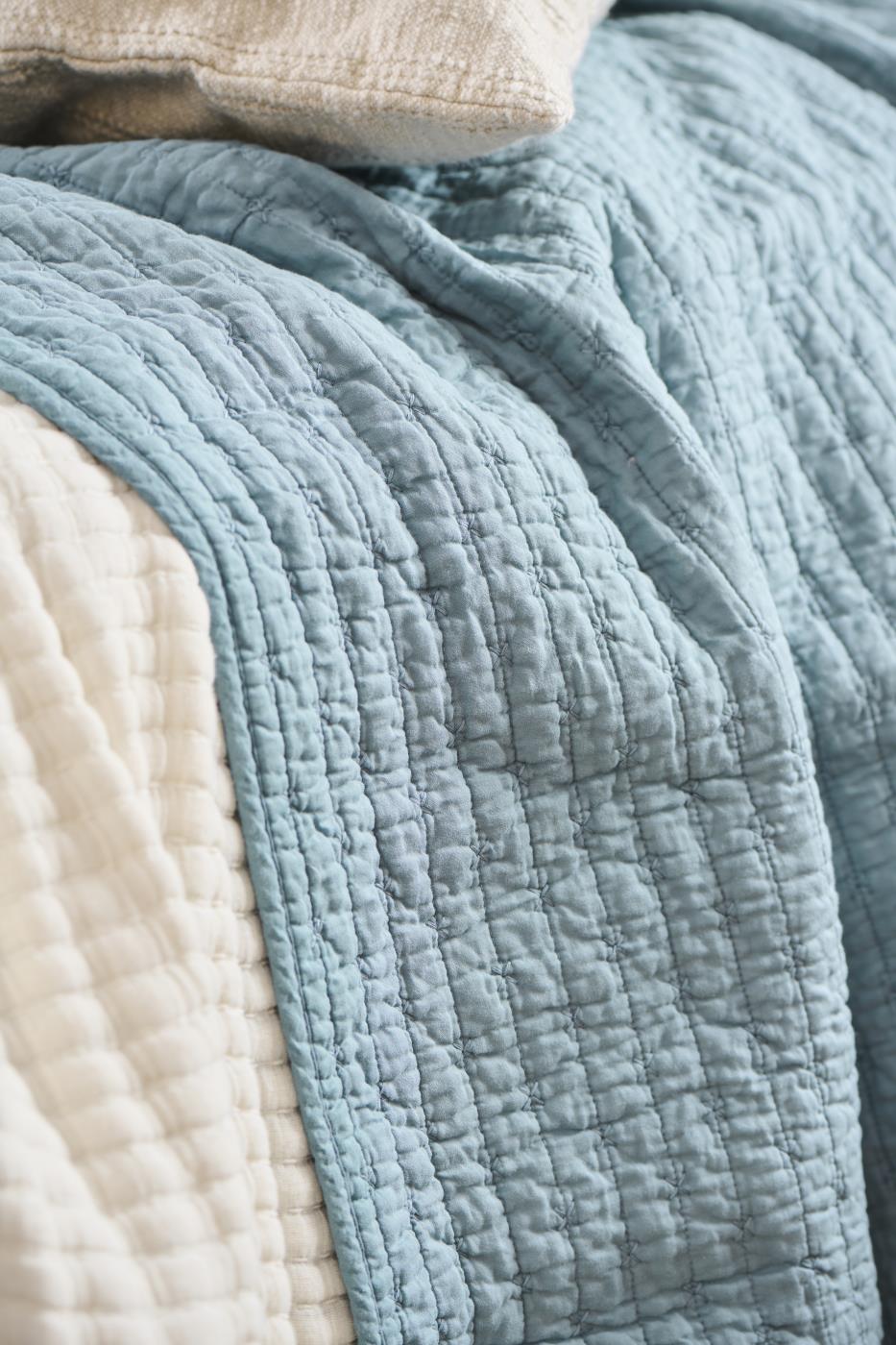 Eine Nahaufnahme eines Bettes mit der IB Laursen Quilt Decke hellblau mit Kreuzstich, drapiert über eine cremefarbene Steppdecke und Kissen, ideal für skandinavische Wohndeko.