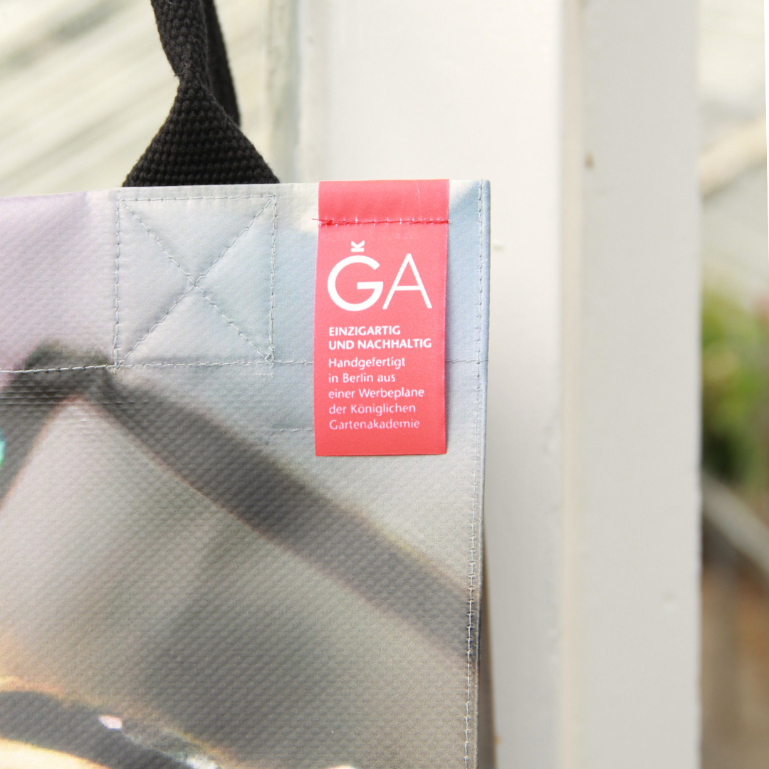 Eine Nahaufnahme der Upcycling KGA Tasche 'Biene' von KGA mit rotem Etikett: Handgefertigt in Berlin aus einem Werbebanner der Königlichen Gartenakademie, hängt sie an einem schwarzen Riemen.