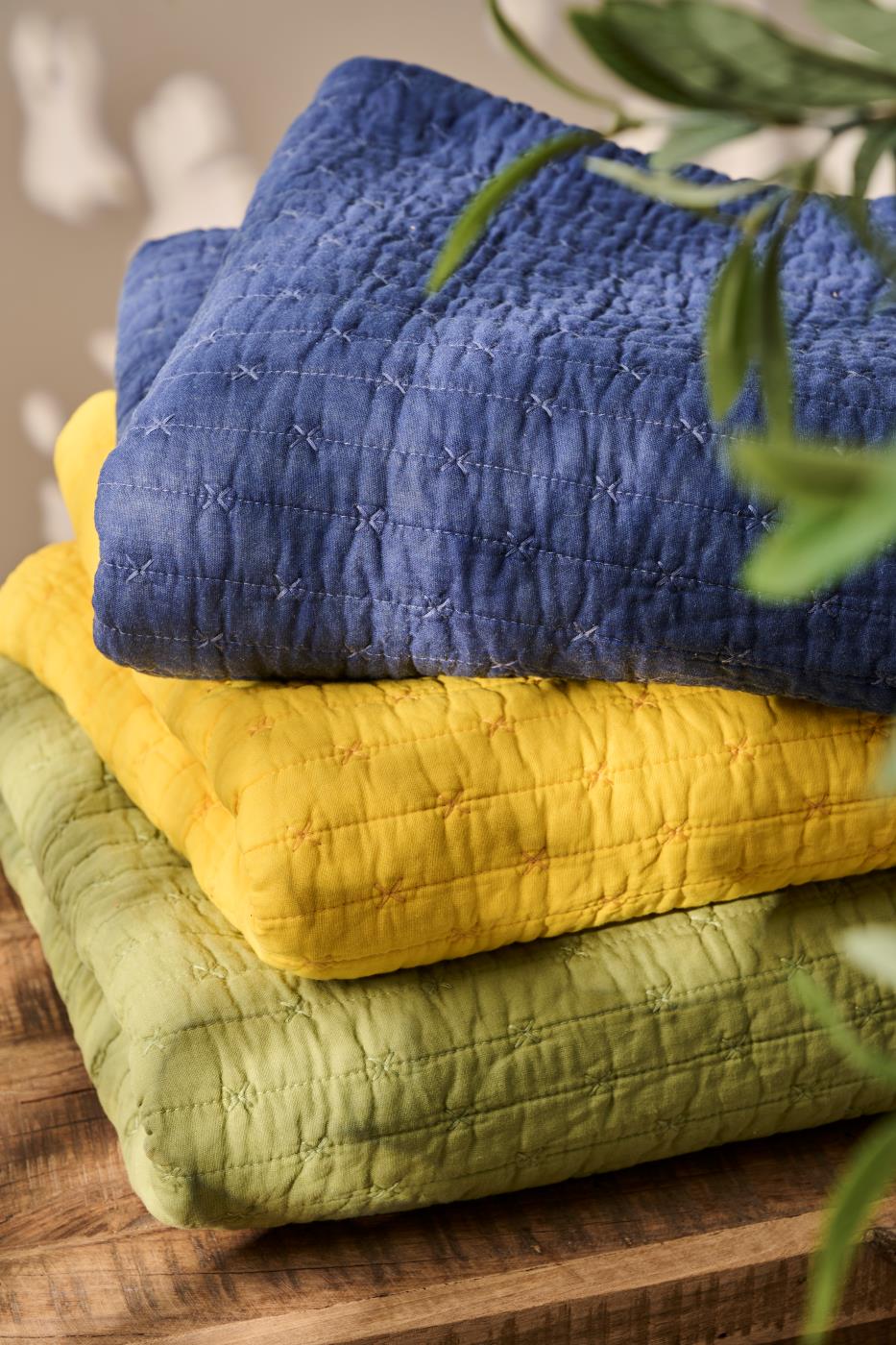 Drei strukturierte Quilts - darunter die IB Laursen Quilt Decke Indigo blau mit Kreuzstich - sind in Blau, Gelb und Grün gefaltet und auf Holz gestapelt, eingerahmt von grünen Blättern vor einem sanft verschwommenen beigen Hintergrund.