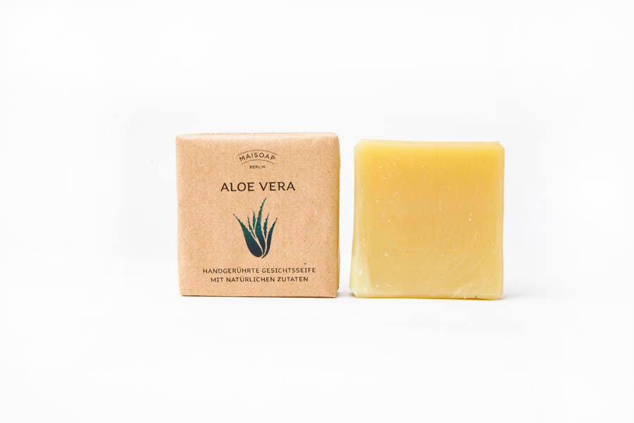 Ein gelber Riegel maisoap berlin - Aloe Vera Seife ruht neben einer braunen Papierverpackung mit einer Illustration der Aloe-Pflanze und einem deutschen Text, ideal für die sanfte Gesichtsreinigung, vor einem weißen Hintergrund.