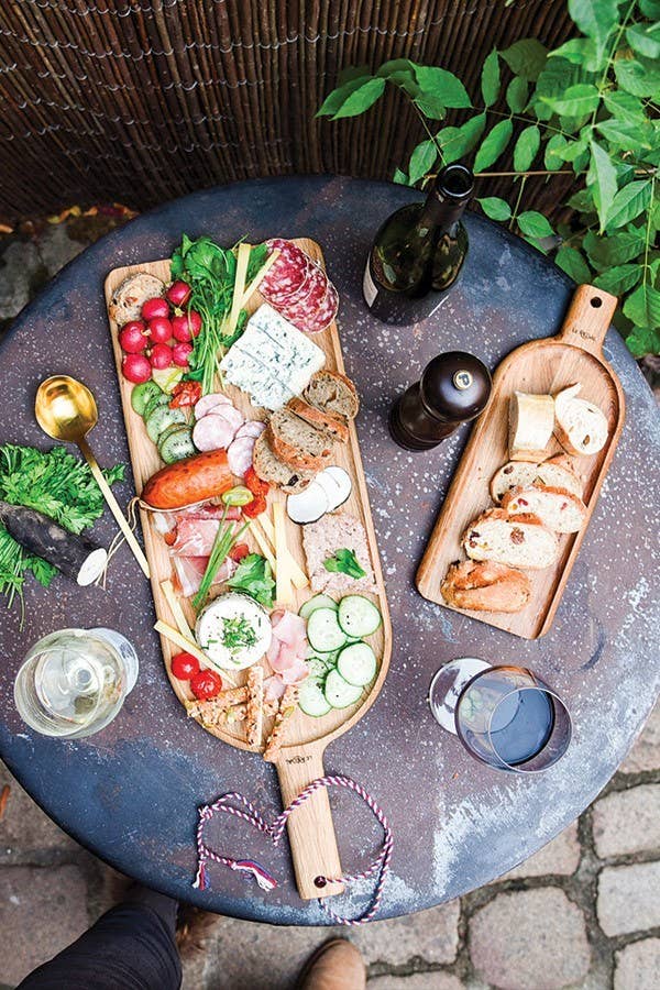 Auf einem runden Tisch im Freien sitzt die LE RÉGAL Große Schaufel, ein Aperitifbrett aus französischem Eichenholz, belegt mit Käse, Fleisch, Brot, Radieschen, Gurken & Grünzeug - ideal für den nachhaltigen Genuss zum Wein.