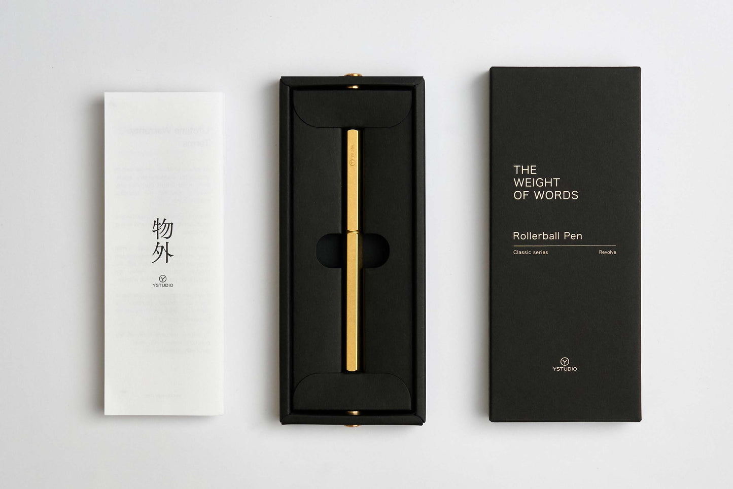 Ein goldener ENTREPÔT - YSTUDIO Klassischer Revolve-Rollerball-Stift (Messing) mit stabilem Griff liegt in einer schwarzen Schachtel zwischen einem weißen japanischen Heft und einem schwarzen Deckel mit der Aufschrift THE WEIGHT OF WORDS. Auf beiden Deckeln ist die Marke ENTREPÔT zu sehen.