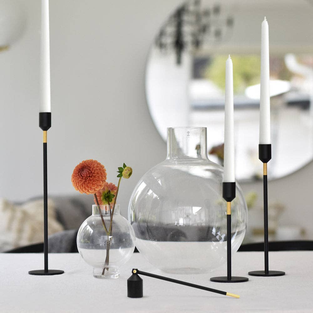 Ein moderner Tisch mit drei Kerzenständern von Solstickan Design – Lichtlöscher – Schwarz, die weiße Kerzen mit metallischen Akzenten halten, zwei runden Glasvasen (eine davon mit einer orangefarbenen Blume) und einem runden Spiegel im Hintergrund.
