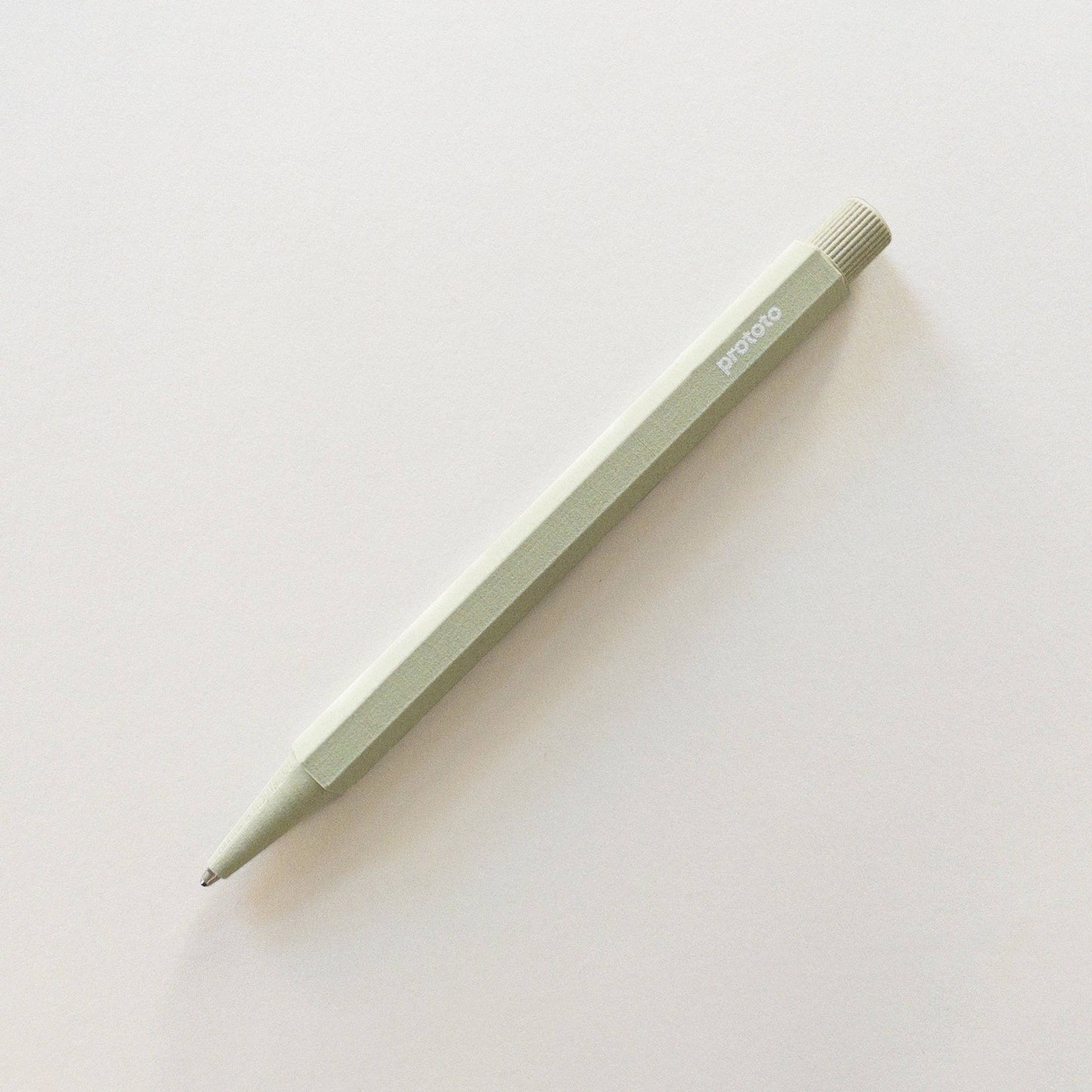 Ein salbeigrüner Prototo Zero Waste Kugelschreiber mit einem sechseckigen Schaft aus klimaneutralem Biokunststoff liegt diagonal auf einer schlichten weißen Oberfläche und trägt den Markennamen Prototo in der Nähe des Radiergummiendes.