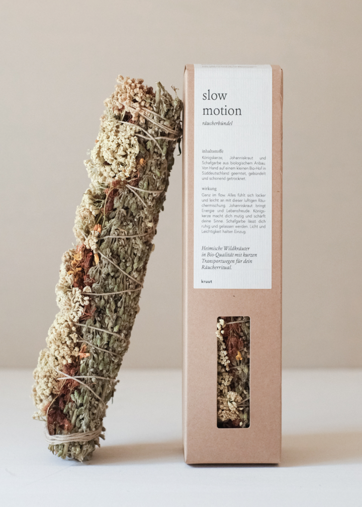 Ein getrocknetes Kräuterbündel von Kruut - Räucherbündel Slow Motion, mit Bindfaden verschnürt, liegt neben einer braunen, rechteckigen Kruut-Dose mit der Aufschrift "Slow Motion", die auf der Vorderseite einen Text und im Inneren ein Fenster mit passenden Kräutern aufweist.