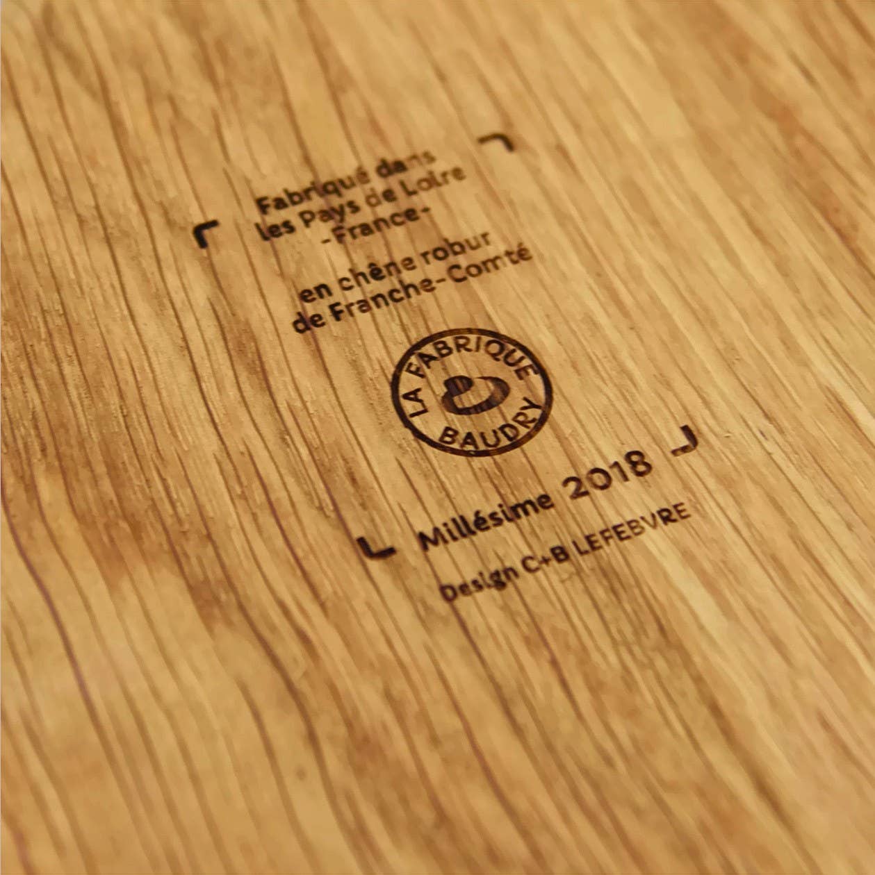 Nahaufnahme der LE RÉGAL - Große Schaufel, ein Aperitifbrett aus französischem Eichenholz, mit eingraviertem französischem Text, dem Logo von La Fabrique Baudry, Millésime 2018, dem Namen des Designers und der charakteristischen Holzmaserung.