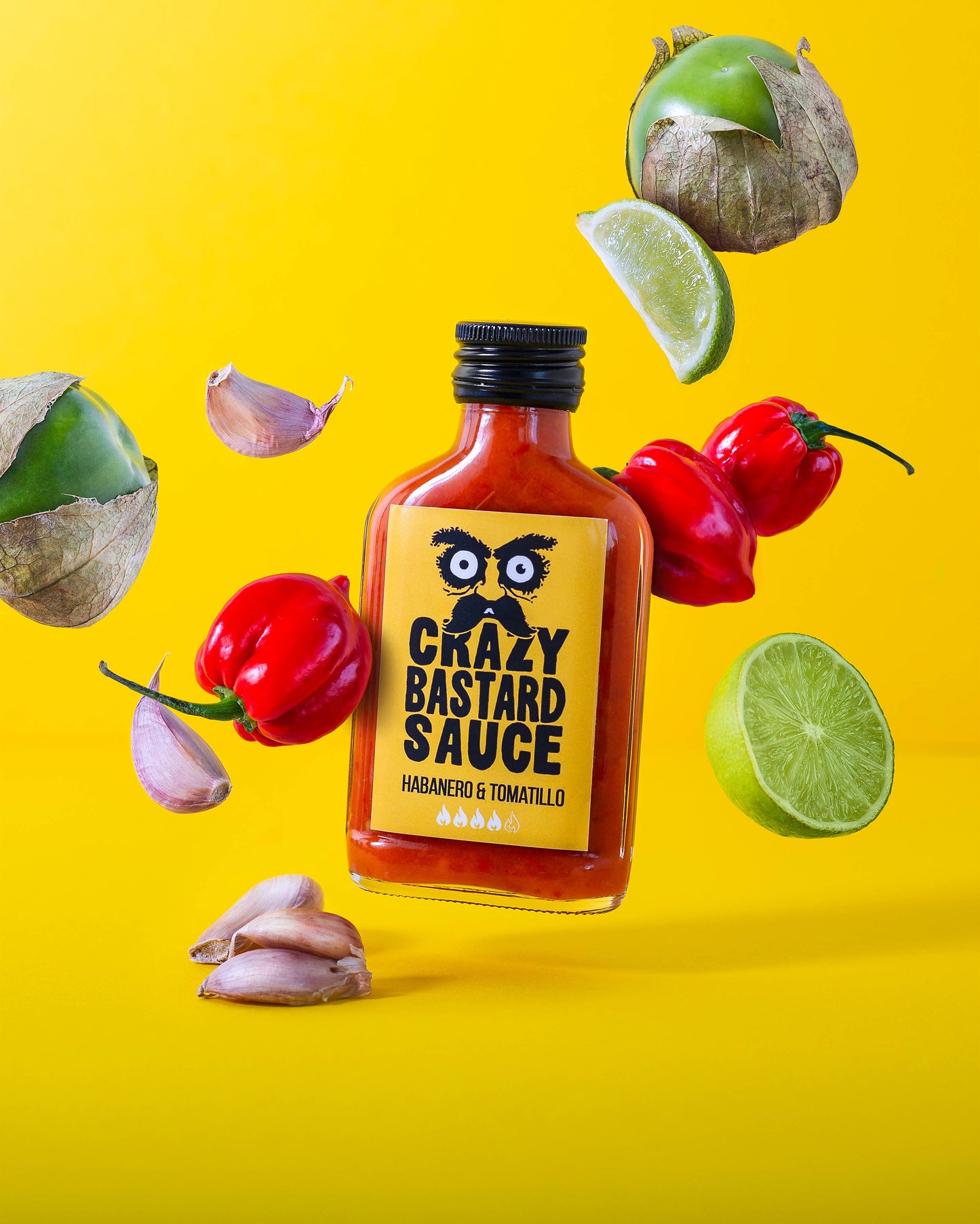 Eine 100 ml Flasche CBS Foods GmbH (Crazy Bastard Sauce) - Habanero & Tomatillo scharfe Soße ist mit Habanero Chilis, Tomatillos, Knoblauchzehen und Limettenhälften auf einem leuchtend gelben Hintergrund abgebildet - ideal für Gewürzliebhaber.