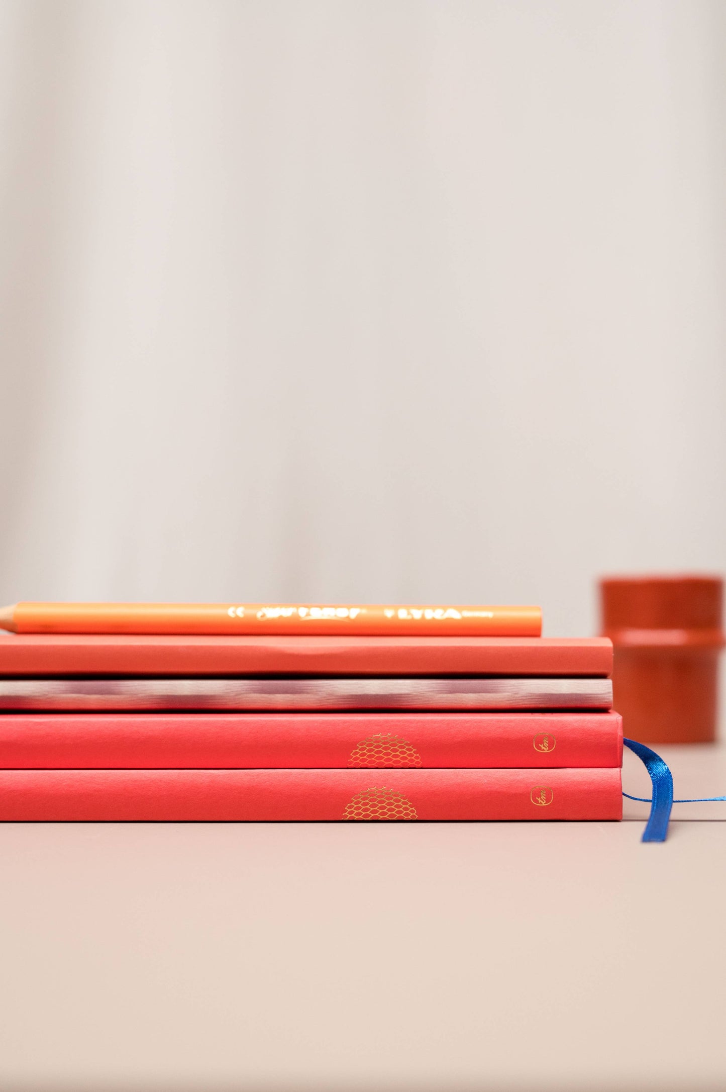Vier "Tinne Mia - Notizbuch mit japanischem Verschluss - Cinnamon" Notizbücher von Tinne Mia sind auf einer hellen Fläche gestapelt, mit einem orangefarbenen Bleistift obenauf und einem verschwommenen orangefarbenen Objekt im Hintergrund.