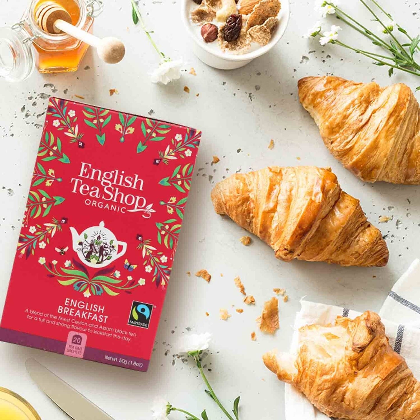 Eine Schachtel English Tea Shop – English Breakfast, BIO Fairtrade, 20 Teebeutel steht auf einer hellen Oberfläche neben drei Croissants, Honig mit Schöpflöffel, einer weißen Blume und Teeblättern – perfekt für eine genussvolle Teezeit.