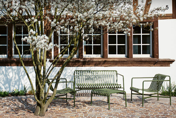 Drei grüne Metallstühle stehen unter einem blühenden Baum mit weißen Blumen, der die Terrassengestaltung aus "Die schönsten Terrassen und Sitzplätze" des Callwey Verlags inspiriert, vor einem weißen Gebäude mit braun gerahmten Fenstern und einer mit Ziegeln gepflasterten Terrasse.