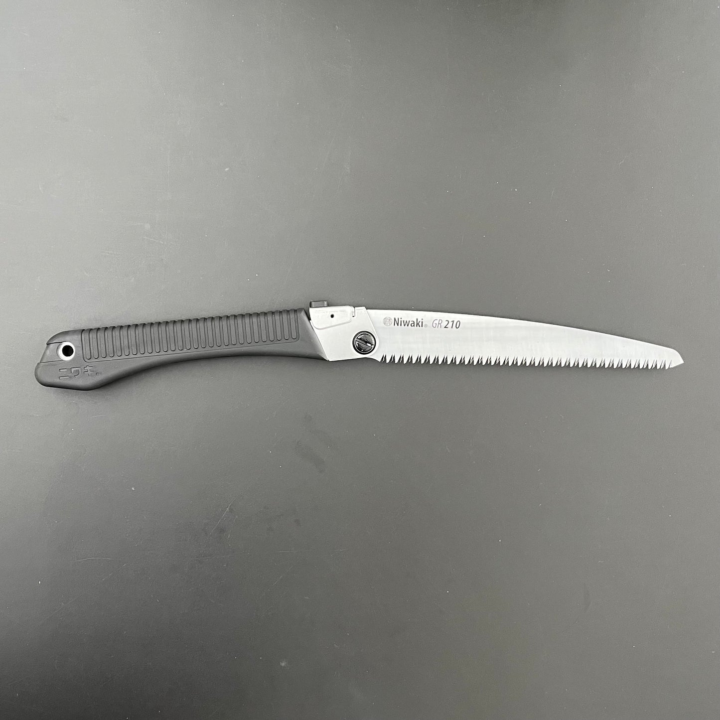 Die Klappsäge Niwaki GR 210 von Niwaki verfügt über einen schwarz strukturierten Griff und eine silberne gezackte Klinge auf einer dunkelgrauen Oberfläche – perfekt für Gartenarbeiten und detaillierte Aufgaben im Freien.