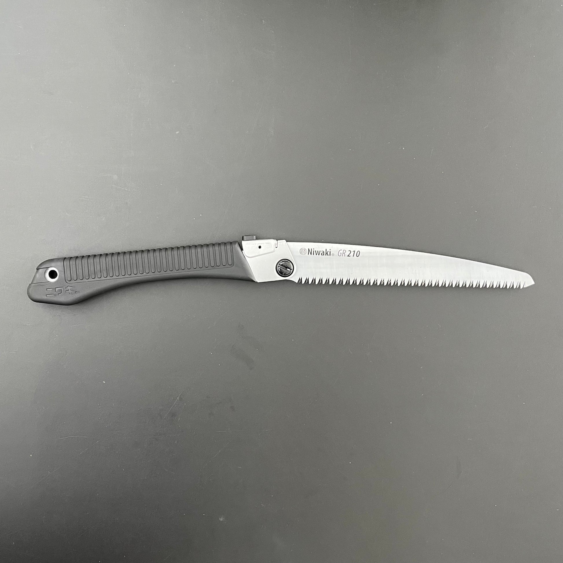 Die Klappsäge Niwaki GR 210 von Niwaki verfügt über einen schwarz strukturierten Griff und eine silberne gezackte Klinge auf einer dunkelgrauen Oberfläche – perfekt für Gartenarbeiten und detaillierte Aufgaben im Freien.