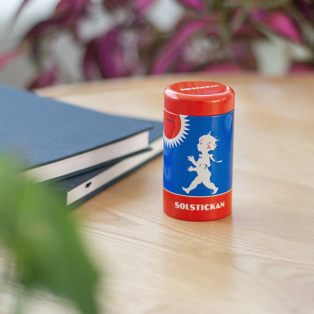Ein Solstickan Design Matches Tube - Originaldesign, in Rot und Blau mit einer weißen Jungensilhouette, ruht auf einem Holztisch neben einem geschlossenen dunklen Notizbuch; verschwommene Pflanzen im Hintergrund erinnern an klassisches schwedisches Design.