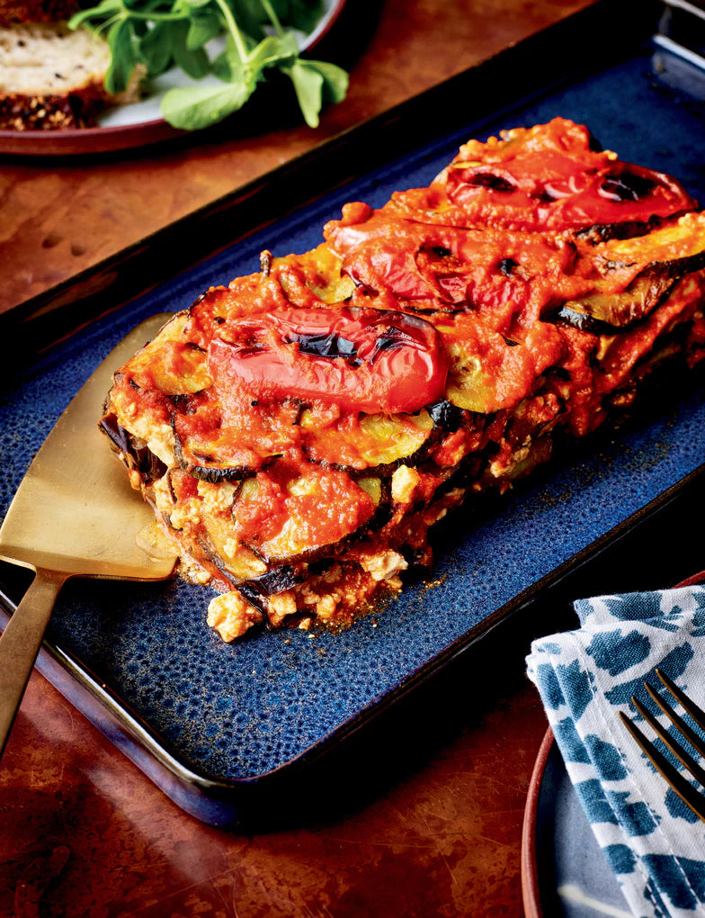 Eine rechteckige gebackene Aubergine mit Parmesan aus der Gewürzküche des Hölker Verlags liegt auf einem dunkelblauen Tablett mit einem goldenen Spachtel. In der Nähe liegen eine Serviette und Grünzeug, das zum Entdecken neuer Rezepte einlädt.
