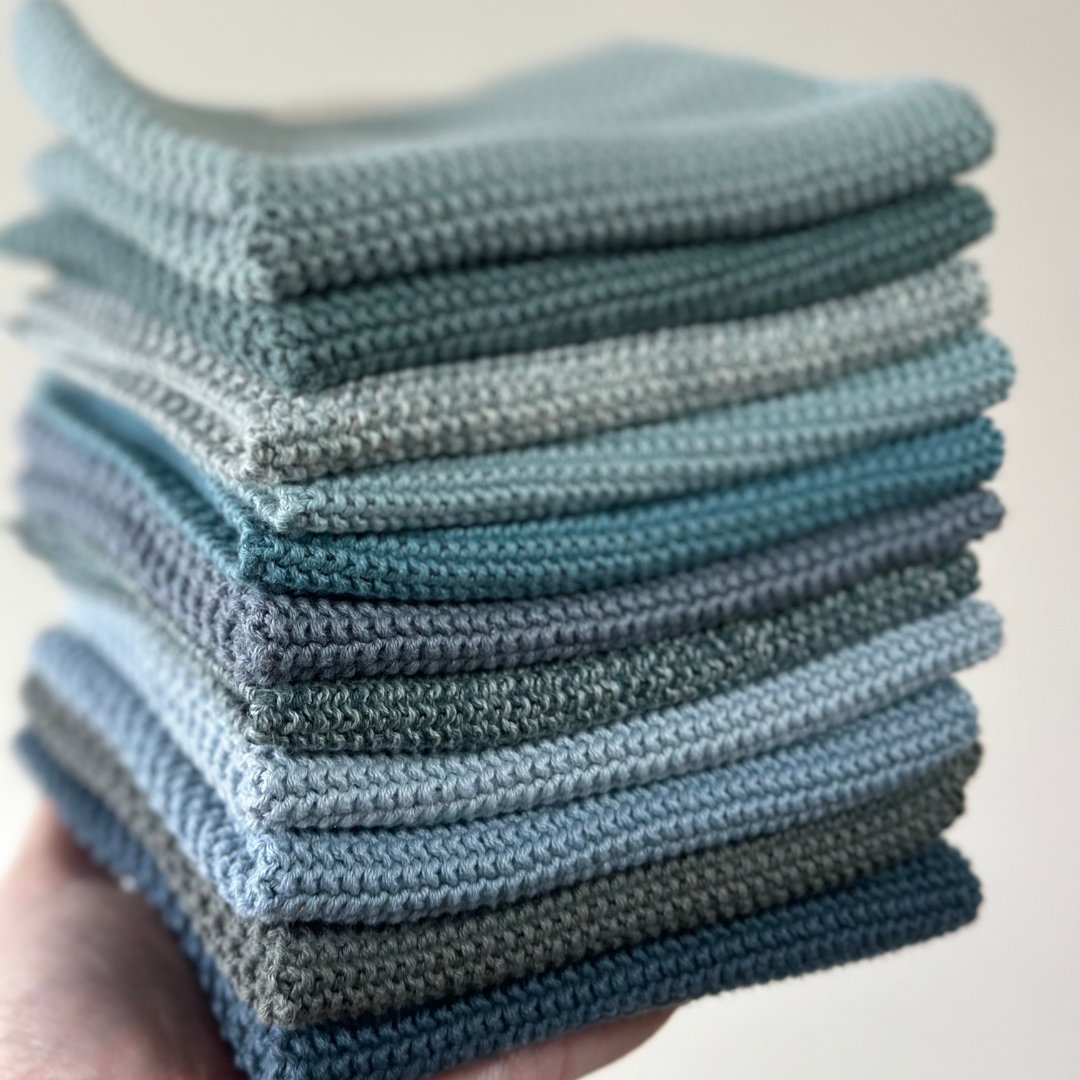 Eine Nahaufnahme einer Hand, die einen ordentlich gestapelten Stapel Aspegren Denmark Spültücher – gestrickt, einfarbig in Blau – hält. Die Tücher sind gleichmäßig gefaltet, strukturiert und umweltfreundlich.