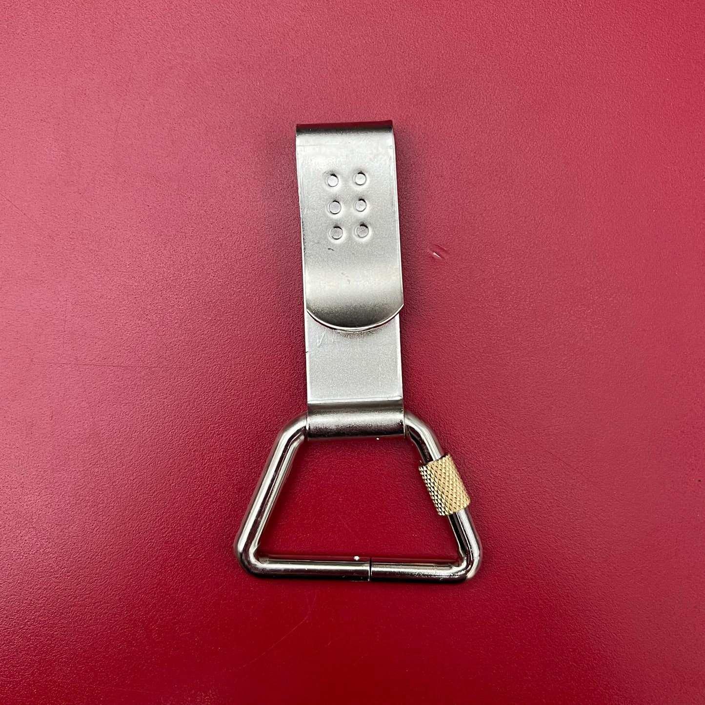Der Niwaki Belt Clip – ein Gürtelclip aus Metall mit dreieckigem Karabiner und goldenem Schraubverschluss – eignet sich ideal für Ihre Gartenschere oder zur Kombination mit dem Niwaki Single Holster, hier auf einer roten Fläche abgebildet.