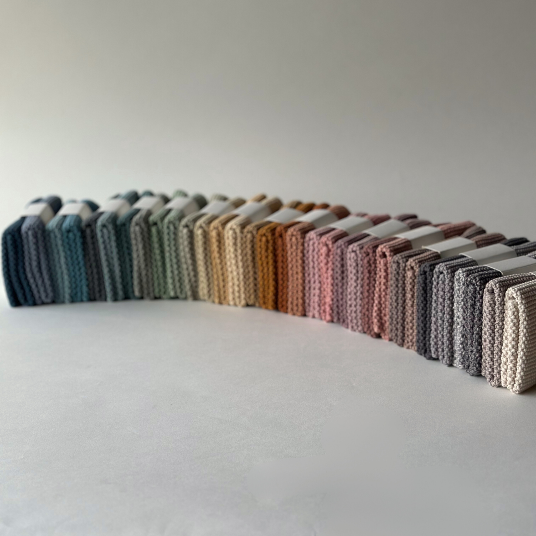 Eine Reihe von Aspegren Denmark Spültüchern, gestrickt aus Bio-Baumwolle in verschiedenen dezenten Farbtönen von Blau und Grün bis Rosa, Grau und Beige, ordentlich nebeneinander auf schlichtem Hintergrund angeordnet.