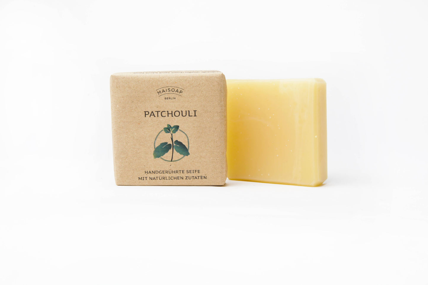Ein gelber Riegel "Patchouli Seife" von maisoap berlin liegt neben der braunen Papierverpackung mit der Aufschrift "Patchouli & Zitronengras", die eine einfache Patchouli-Blatt-Illustration und einen deutschen Text auf weißem Hintergrund zeigt.