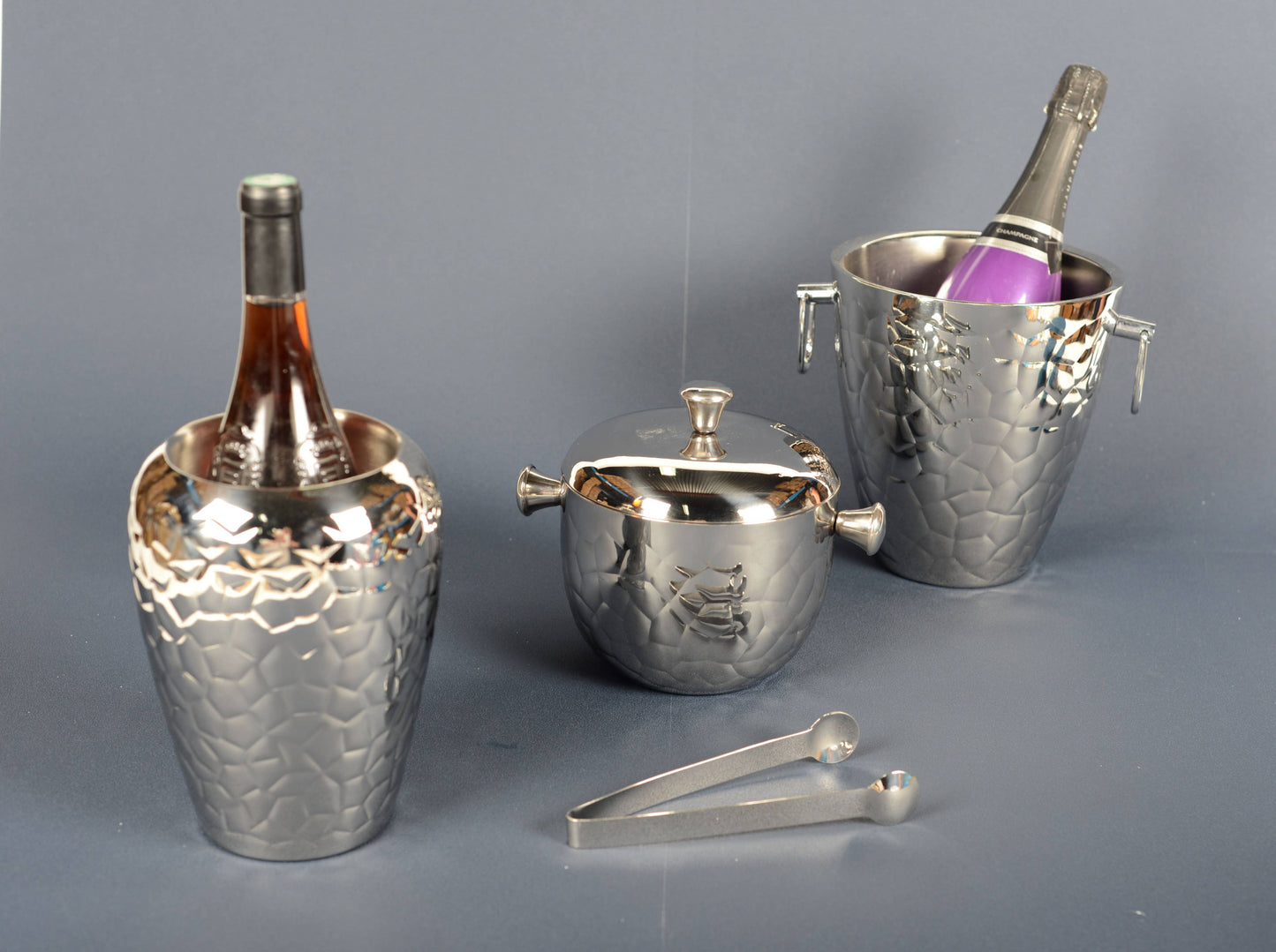 Zwei doppelwandige AULICA Weinkühler in Silber (13 cm hoch) halten Weinflaschen, während ein AULICA Eiskübel mit Deckel und Griffen daneben steht. Davor befindet sich eine Eiszange aus Metall, alles angeordnet auf einer grauen Fläche mit grauem Hintergrund.