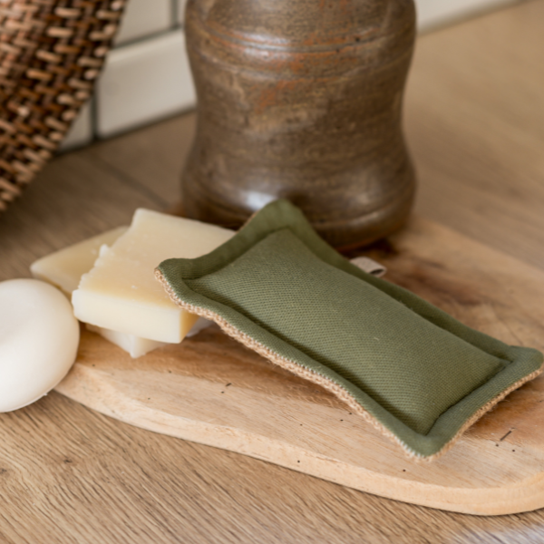 Der „Simple Things - Waschbarer Schwamm | Die Scharfen“ von Simple Things, handgefertigt in Frankreich aus Baumwolle/Jute mit hypoallergenem Futter, liegt auf einem Holzbrett neben Seifenstücken und einem Keramikbehälter.