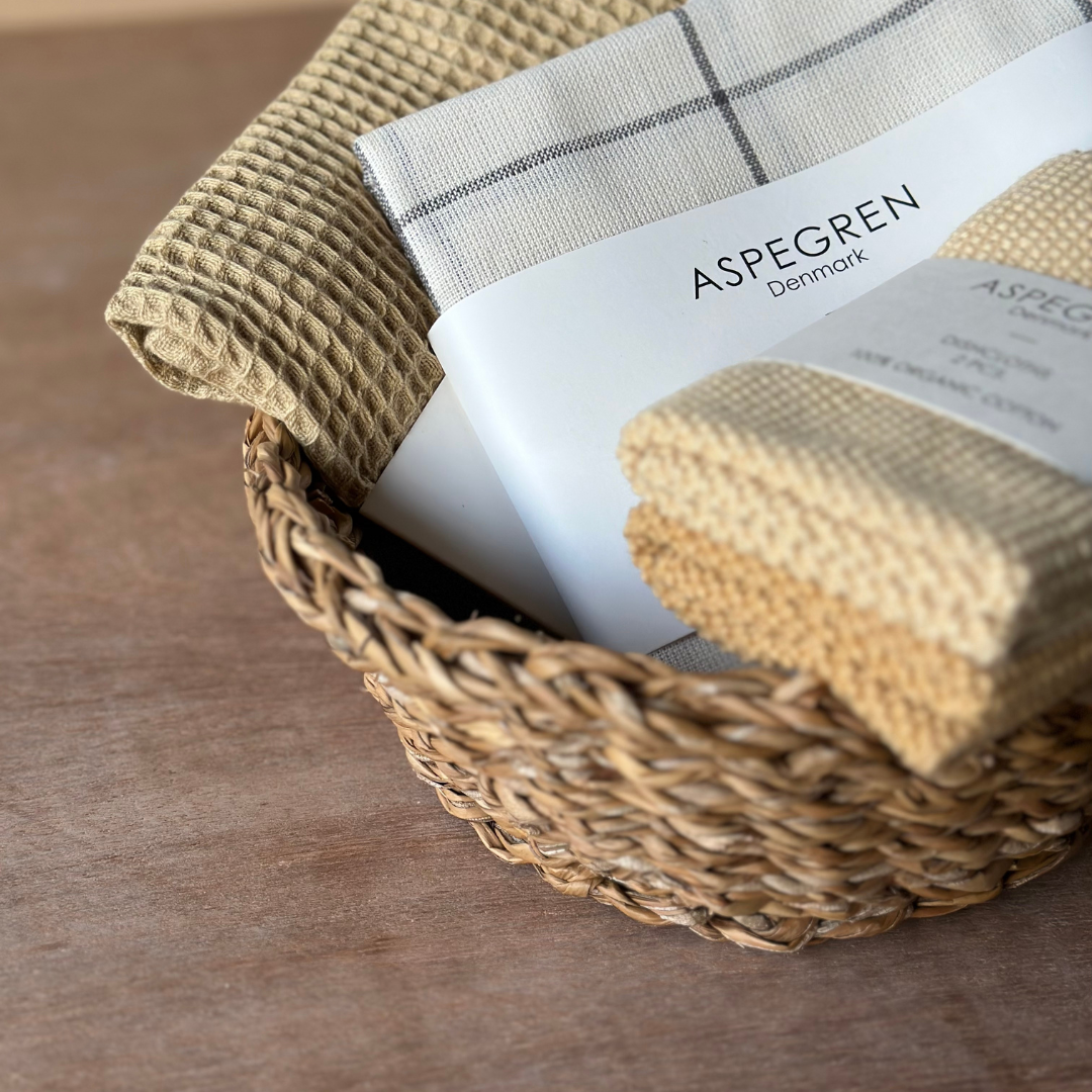 Ein geflochtener Korb enthält goldfarbene, gestrickte "Spültücher" von Aspegren Denmark mit einfarbigen Motiven und Markenverpackungen, die auf einer Holzfläche ausgestellt sind.