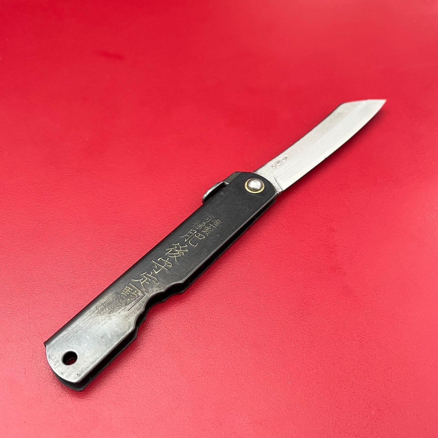 Ein kompaktes Niwaki SK Higonokami Klappmesser von Niwaki mit schwarzem Griff und japanischen Schriftzeichen liegt auf einer glatten, leuchtend roten Oberfläche. Die scharfe Klinge ist teilweise geöffnet und nach rechts abgewinkelt.