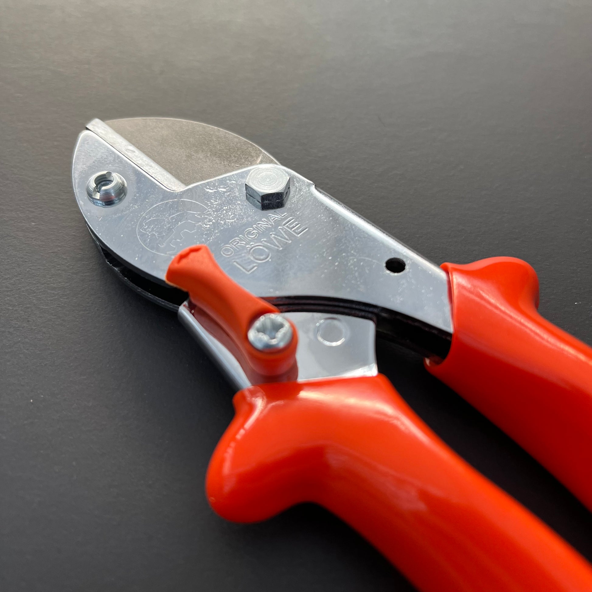 Eine Nahaufnahme der Original Löwe 5 - Classic Easy Cut Gartenschere mit Amboss-Technik und leuchtend orangefarbenen Griffen, platziert auf einer dunklen Fläche; die Klinge ist mit dem Markennamen graviert.