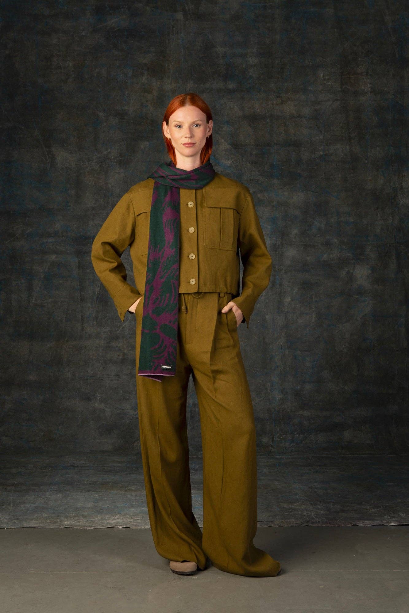 Eine rothaarige Frau steht vor einem dunklen Hintergrund, trägt einen lockeren olivgrünen Hosenanzug und den Wolvis Schal aus Merinowolle - Cerura Traube midi. Ihre Hände stecken in den Taschen und sie hat einen ruhigen Gesichtsausdruck.