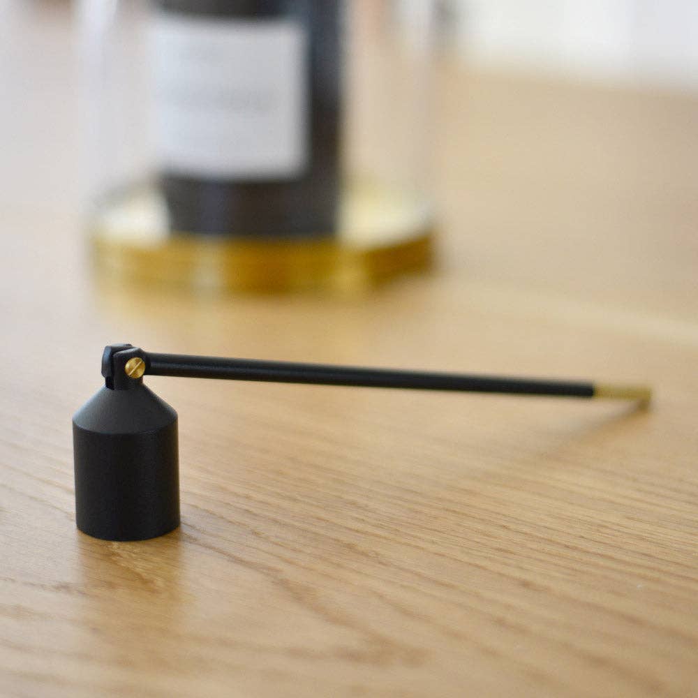 Der Solstickan Design - Lichtlöscher - Schwarz, ein schwarzer Kerzenlöscher aus Metall mit langem Griff von Solstickan Design, ruht auf Holz mit einer verschwommenen Kerze und einem Glasobjekt im Hintergrund.