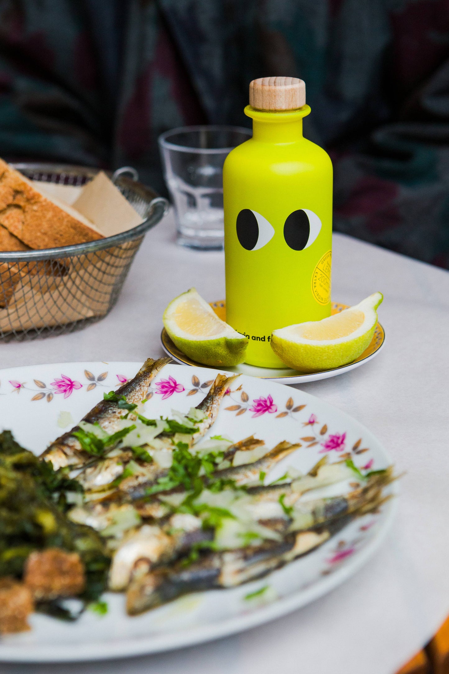 Ein Teller mit gegrillten Sardinen mit Kräutern, Grünzeug und Zitronenspalten steht neben einer leuchtend gelben Flasche yiayia and friends - Natives Olivenöl extra mit Zitrone, einem Brotkorb und einem Glas Wasser.