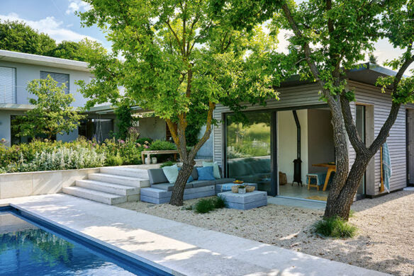 Ein modernes Gästehaus am Pool mit Glasschiebetüren wird von Bäumen beschattet. In der Nähe bietet ein gepolsterter Sitzplatz auf Kies eine perfekte Terrassengestaltung, inspiriert von "Die schönsten Terrassen und Sitzplätze" aus dem Callwey Verlag. Eine Treppe führt in das darüber liegende Haupthaus.